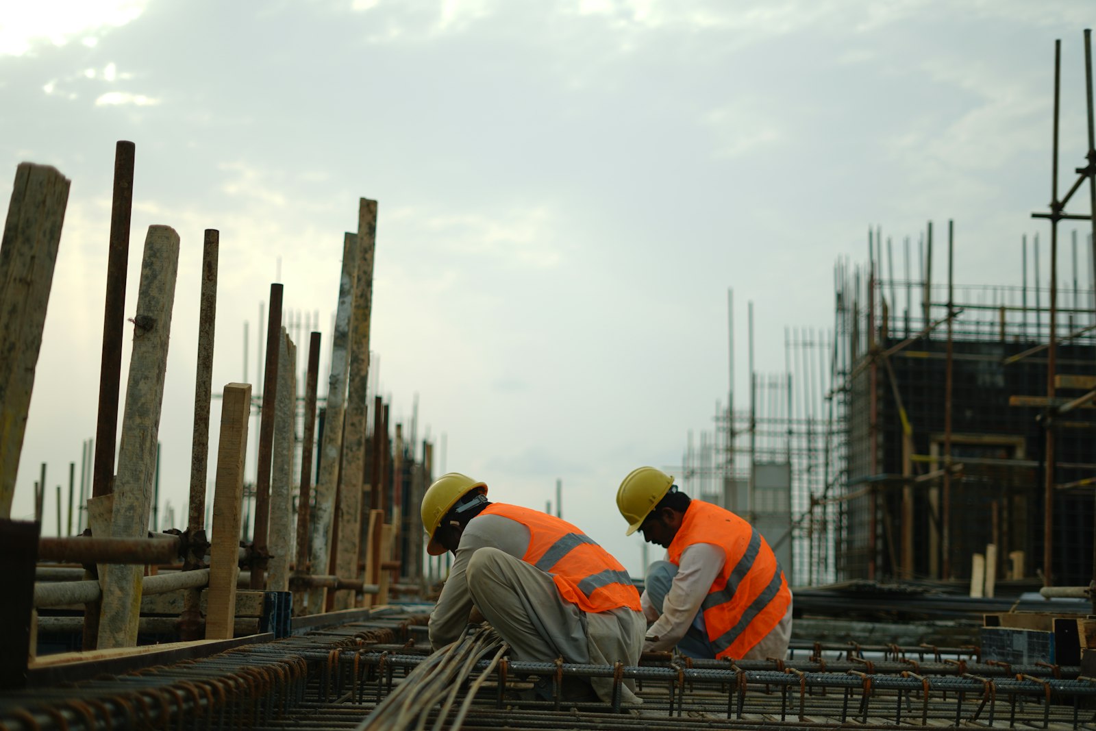 Two construction workers in hard hats and safety vests tying rebar on a building site.