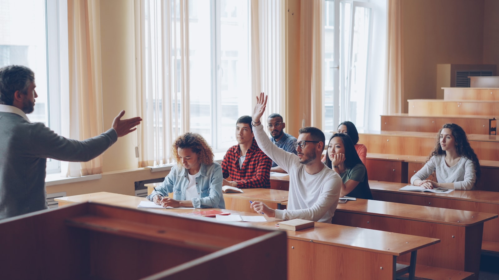 A university student raising a hand to ask a question in a sunlit lecture room.