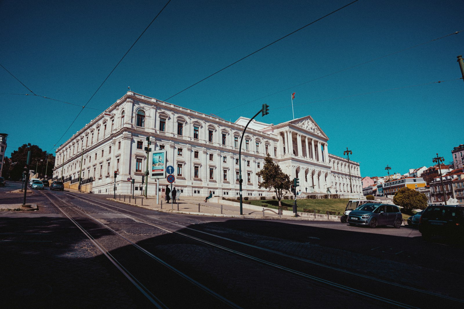 A grand white colonnaded government building with a flag on its roof, seen from a city street under a clear blue sky.