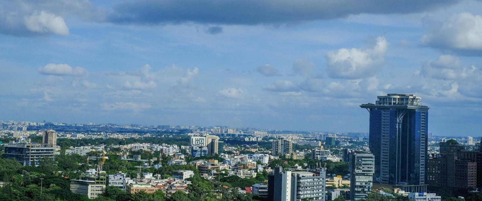 A green, tree-covered city skyline with a tall modern glass skyscraper, under a partly cloudy blue sky.